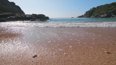 Close-up-ocean-clear-water-waves-and-sea-foam-rolling-into-a-sandy-beach-in-Costa-brava,-Spain