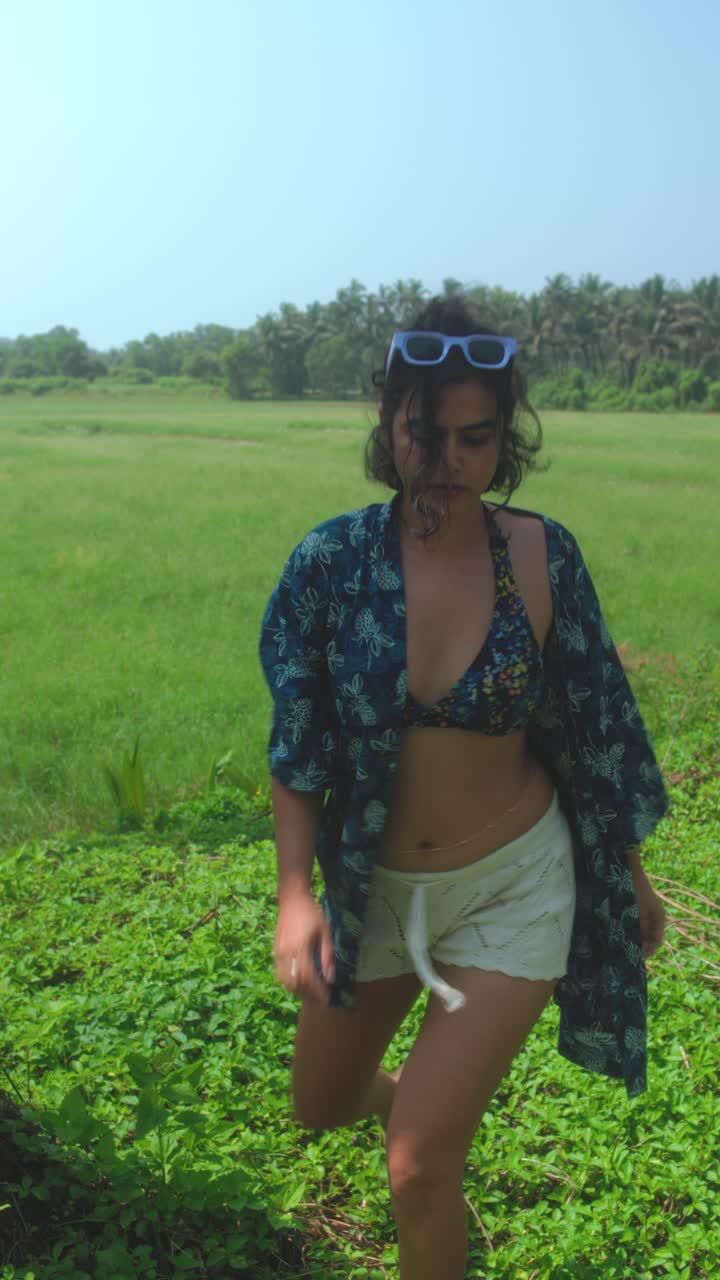 A carefree girl in a bikini top, kaftan, and sunglasses steps out of a lush green field toward the camera. This vertical video embodies summer vacation, freedom, and a tropical nature lifestyle