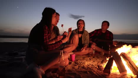 Accelerated-footage-of-friends-sitting-on-the-beach-on-a-plaid-near-the-bonfire,-frying-food-with-wooden-sticks,-taking,-talking