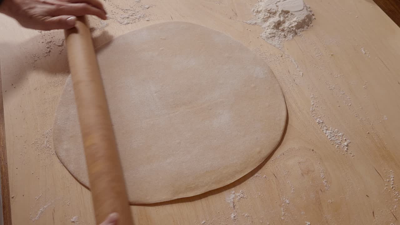Mani di una persona anziana che usano un mattarello di legno per stendere l'impasto su una superficie infarinata, preparando pasta fatta in casa o prodotti da forno.