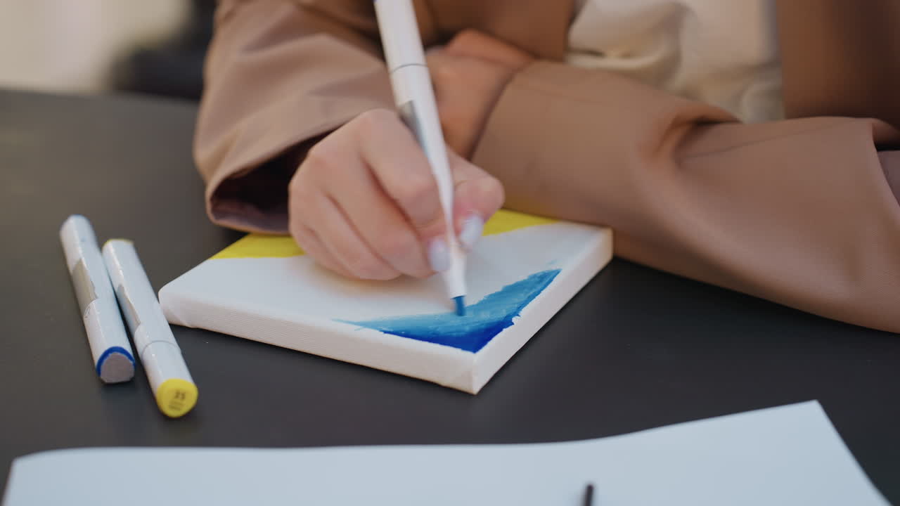 female artist decorating small surface with colorful markers, creative young lady using markers to bring bright colors to miniature painting at shared table in lively public setting