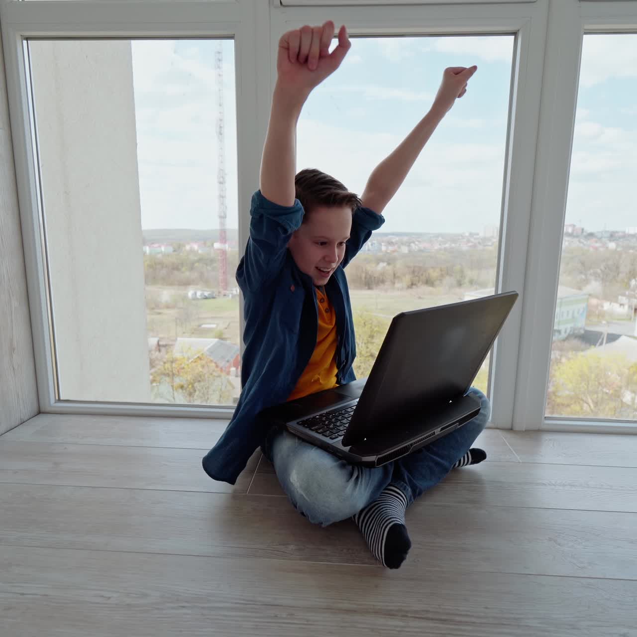 Teen boy sitting on the floor by the window and playing on laptop. Boy plays video games and celebrates victory. Gaming at home.