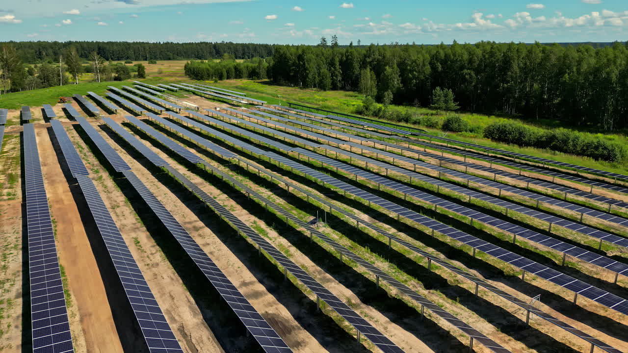 Aerial rearview perspective of a large solar farm construction site with rows of panels in a green landscape, orbit along rows