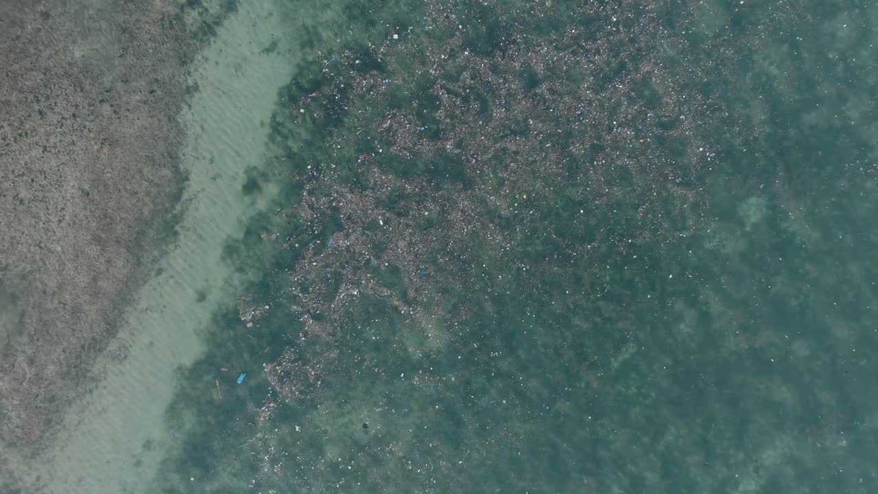 Top down quickly ascending drone shot of polluted water filled with trash floating over dead coral reef in the turqouise tropical water of Bali Indonesia