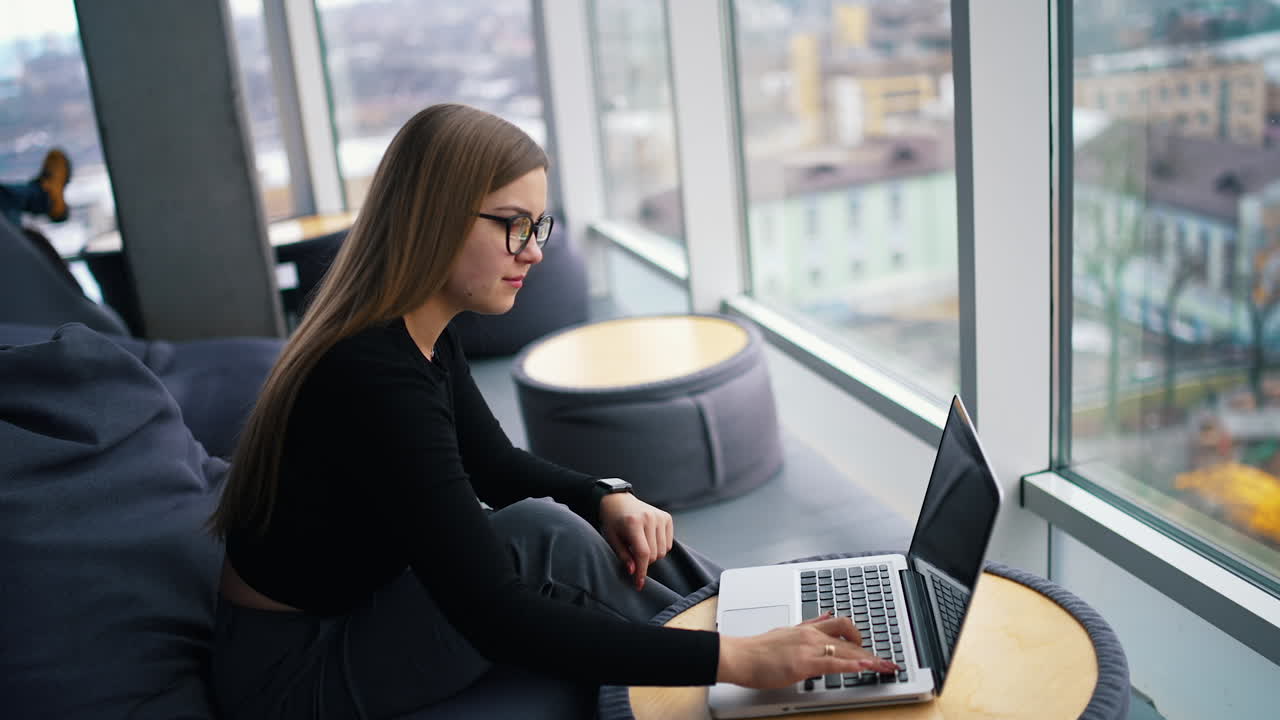Stylish young woman in office interior. Beautiful business woman in glasses working on laptop computer near the window city view.