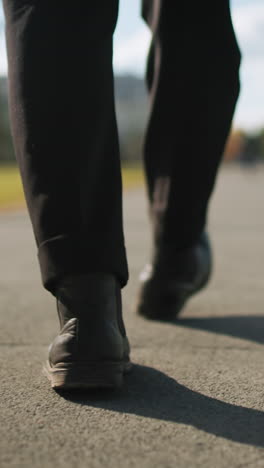 leg view of individual in black trousers and shoes walking confidently outdoors on tarred road with shadow cast on ground, surrounded by urban trees, modern buildings, and sunny blue sky