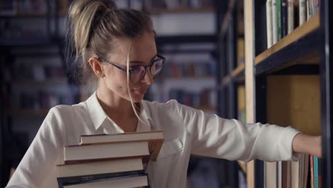 Attractive-woman-holding-pile-of-books-in-the-library,-looking-for-another