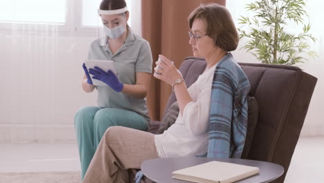 senior woman drinking water while talking to female doctor in medical mask and protective screen using a table