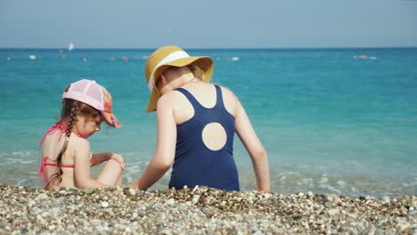 Dos-Chicas-Están-Sentadas-En-Un-Guijarro-Junto-Al-Mar-En-Busca-De-Hermosas-Vacaciones-De-Guijarros-Con-Niños-En