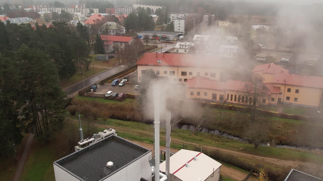 An aerial view of a small industrial plant with chimneys emitting steam or smoke near a residential neighborhood surrounded by trees