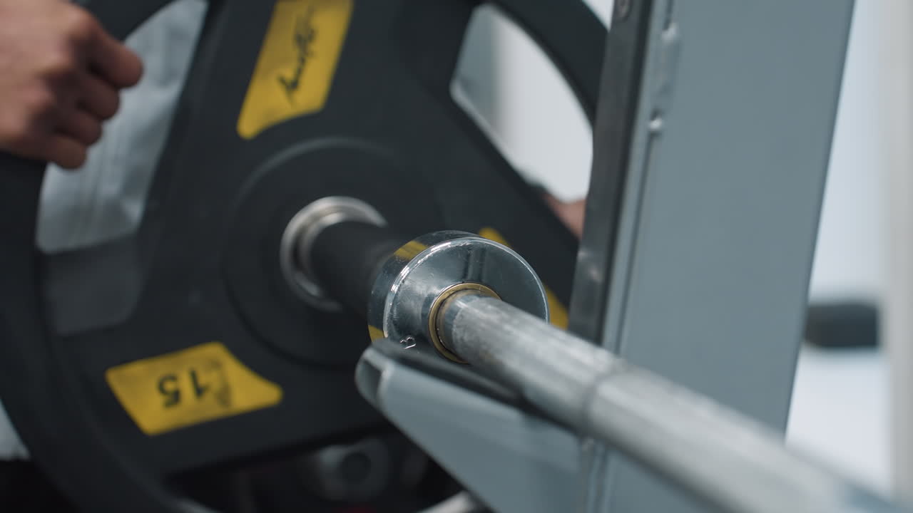 Close up of person returning weight plate onto barbell rack, showing hands guiding heavy iron plate into position on chrome barbell sleeve on rubber flooring in gym environment after workout session