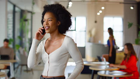 Mujer-De-Negocios-Sonriente-Parada-En-Una-Concurrida-Oficina-Abierta-Hablando-Por-Teléfono-Móvil