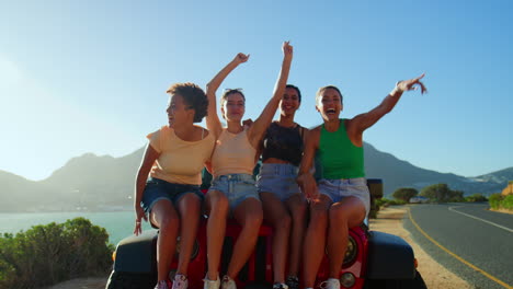 Portrait-Of-Laughing-Female-Friends-Sitting-On-Hood-Of-Open-Top-Car-On-Road-Trip