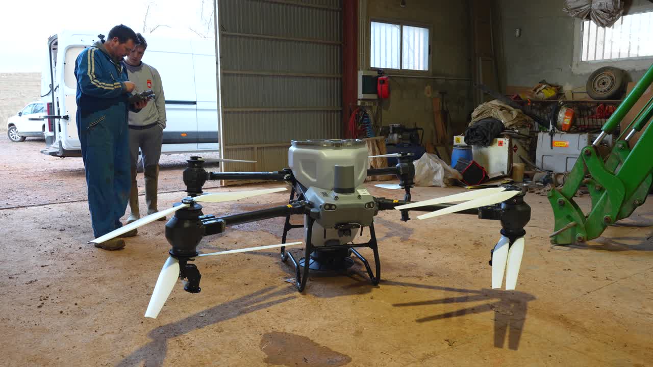 Un agriculteur expérimenté instruit un homme plus jeune à l'intérieur d'une grange sur la configuration d'un drone agricole à l'aide de la télécommande