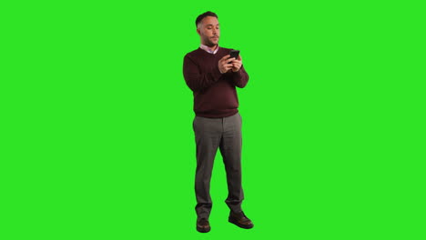 Full-Length-Studio-Portrait-Of-Mature-Male-Teacher-Or-Businessman-Standing-Against-Green-Screen-Using-Mobile-Phone-To-Message-Or-Browse-Online