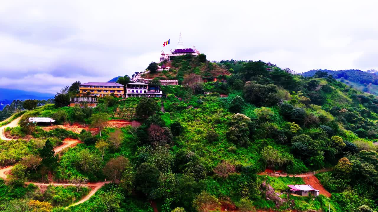 A Sri Lankan Buddhist Asapuwa Temple sits atop a lush hill, surrounded by tropical vegetation, winding paths, scattered rural structures, and panoramic mountain scenery.