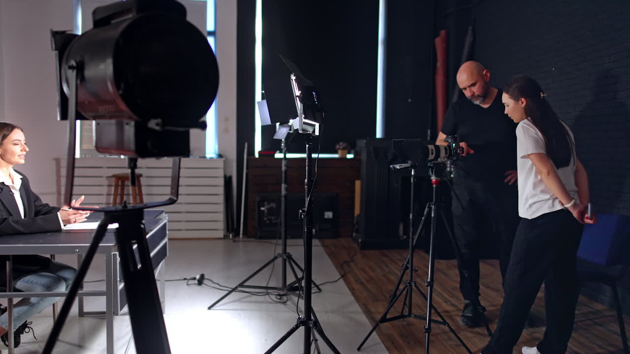 Male photographer and a brunette girl look at the camera. Long-haired woman sitting at desk in the light of soffits talks for the video.