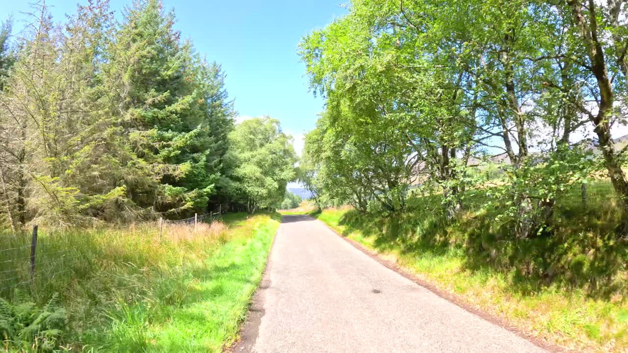 A vehicle travels a narrow country road bordered by grassy hills, trees, and open countryside under bright daylight with clear blue skies