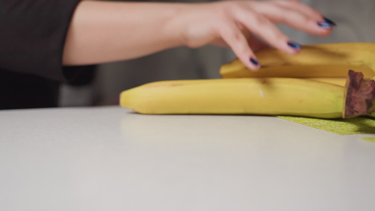 Close up manicured hands carefully lifting banana bunch from table into nylon bag, natural light highlights ripe fruit yellow peel, gesture emphasizes fresh produce, clean surface