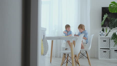 two boys brothers sit at a table in the living room and draw with pencils on paper. mom comes up and hugs the boys, smiling and stroking his head. praises and helps to make the task