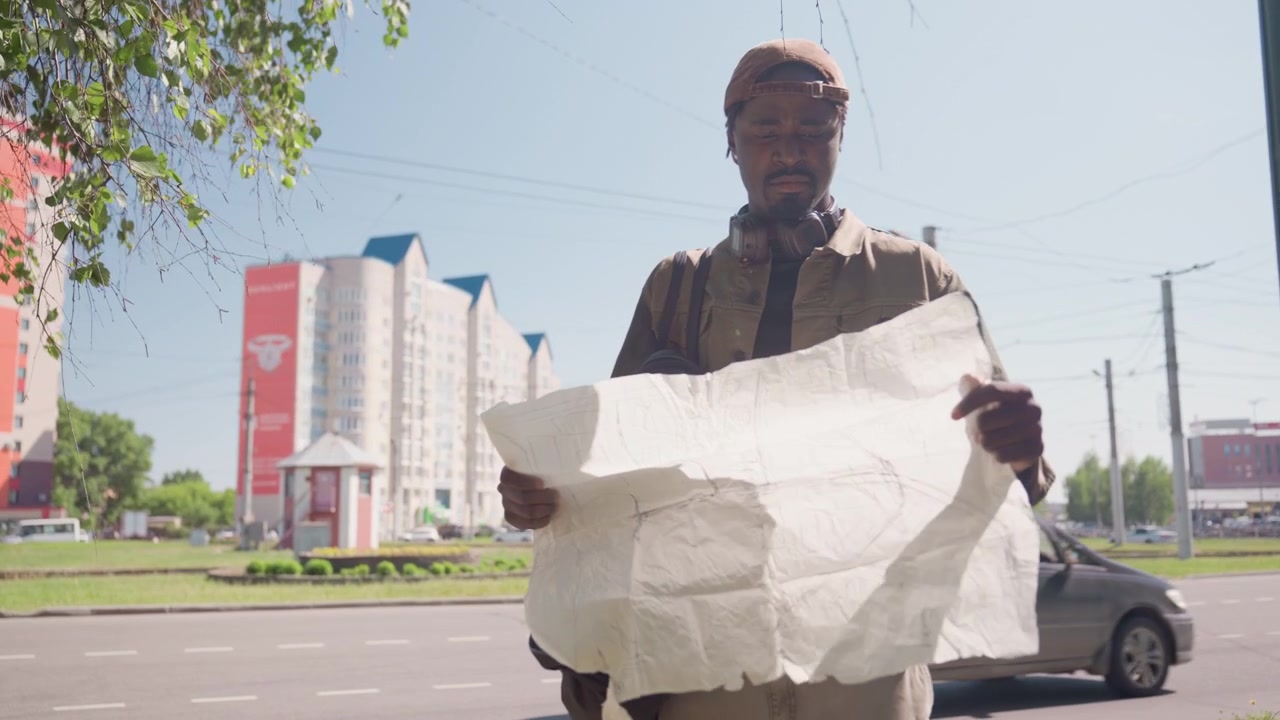 Close up of man holding large white paper and carrying bag on street with cars passing by. Man wears cap headphones around neck tall buildings visible in background under clear sky