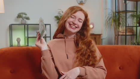 successful rich young caucasian woman holding dollar bill money fan sitting on sofa couch at home