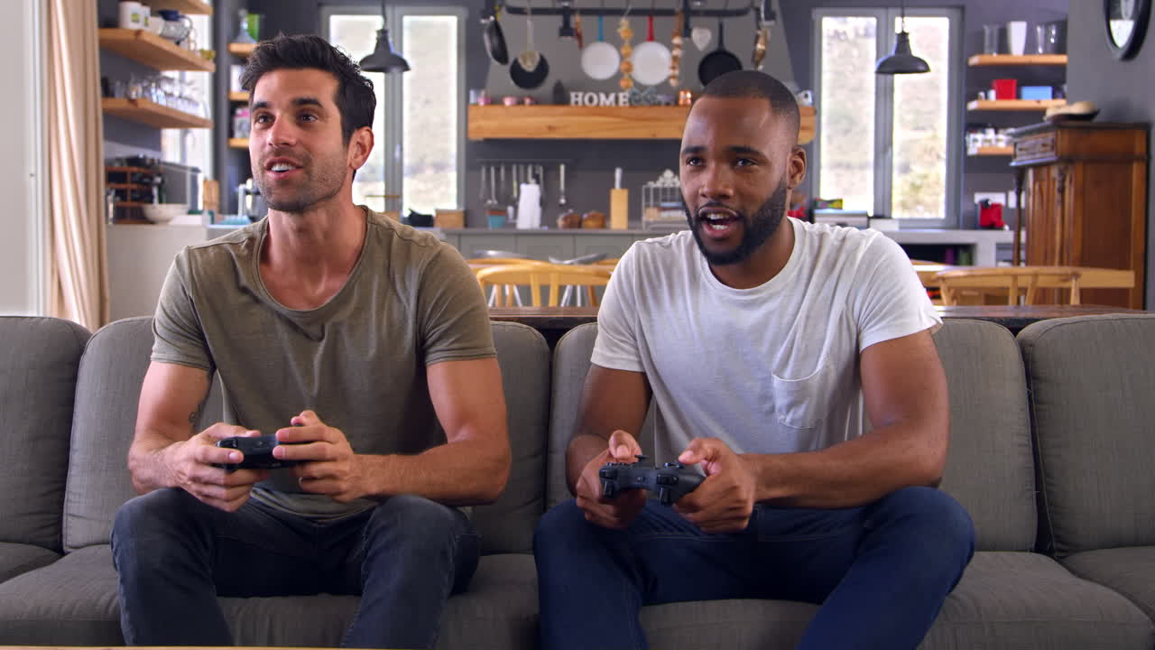 Two Male Friends Sitting On Sofa In Lounge Playing Video Game