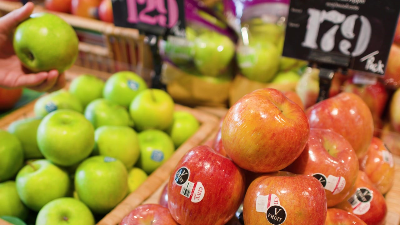 En persons hånd plukker et grønt eple fra en supermarkedhylle fylt med frisk frukt