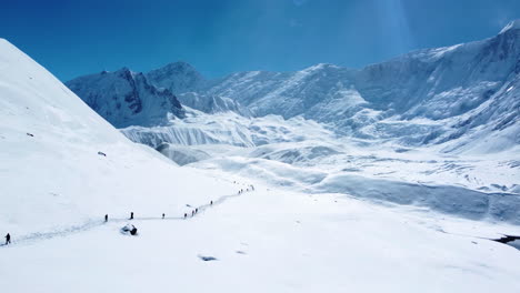 Drone-shot-of-mesmerizing-view-on-snowy-land-surrounded-by-mountains-and-people-walking-in-long-trail-midday-sunlight