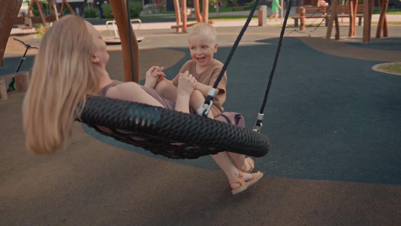 Donna e giovane ragazzo condividono un momento gioioso su un'altalena a pneumatico in un parco giochi all'aperto. Il bambino siede dietro l'adulto, tenendosi per mano mentre ondeggiano dolcemente insieme sotto la piena luce del giorno. Elementi circostanti