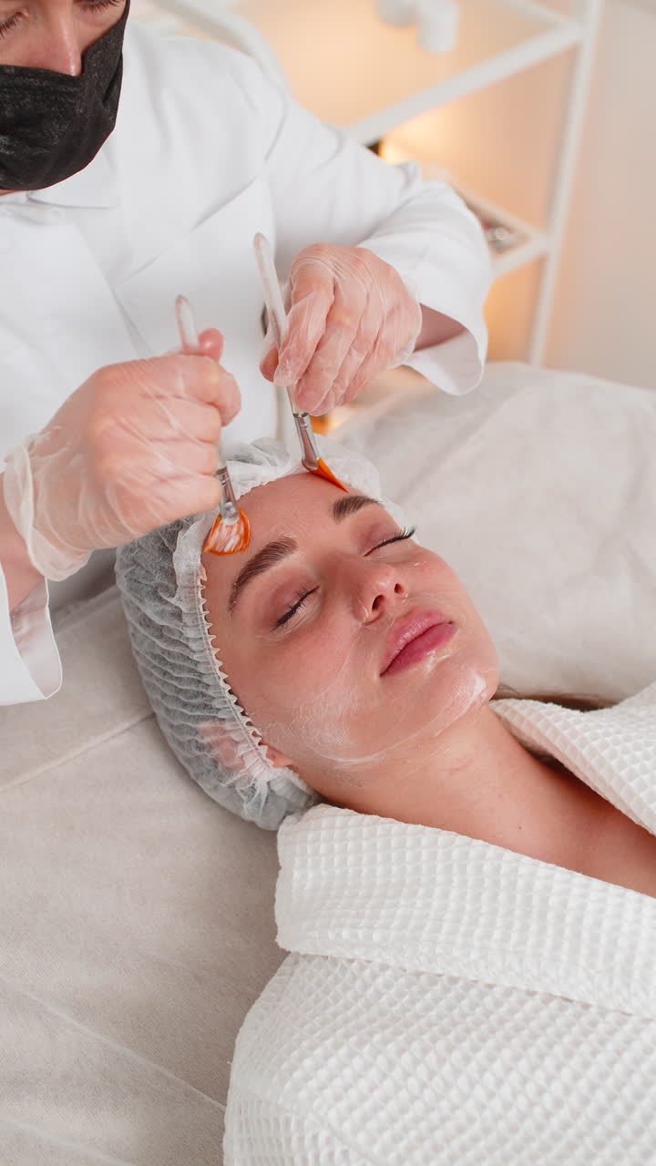 Esteticista aplicando crema médica hidratante con cepillos en el rostro de una mujer de mediana edad durante un tratamiento enzimático
