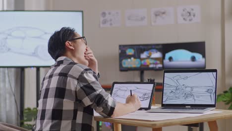 side view of asian male yawning while drawing new car design concept on a tablet in the studio with tv and computers display 3d electric car model