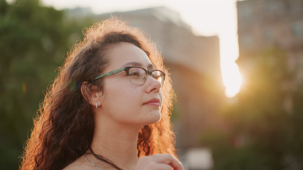 Mujer con vestido rojo en una azotea durante el atardecer, giro de cabeza confiado y movimiento de cabello, gafas reflejando el brillo de la ciudad, blogger de estilo de vida mostrando un look, viajera haciendo una pausa para ver el horizonte, ambiente alegre con lente cálida.