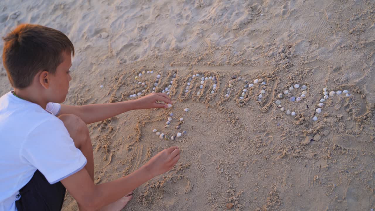 Boy is putting with pebbles the word summer on a sand beach. Child laying the smiley sign with small shells on sand background. Happy childhood.