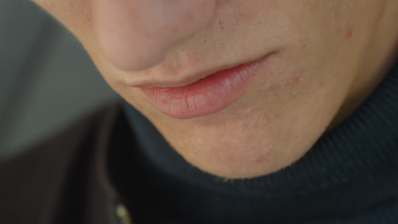 Primo piano della bocca e delle labbra di un uomo caucasico. Parte inferiore del viso illuminata delicatamente con leggera barba incolta e acne lieve, movimento labiale impercettibile che suggerisce un sussurro e una prova, espressione pensierosa catturata in un intimo studio al chiuso