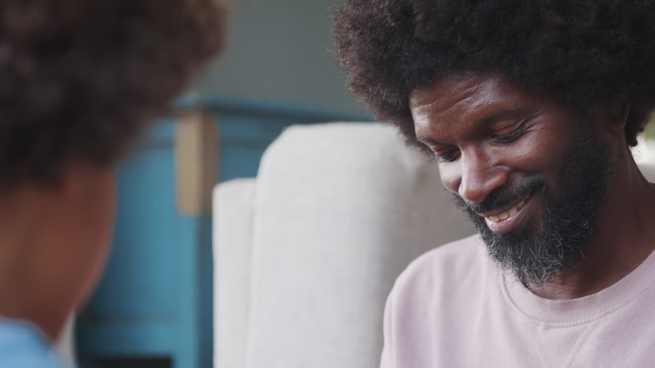 Close up of middle aged black father sitting opposite his son at home talking, close up, head and shoulders, selective focus