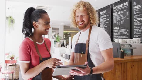 happy diverse male and female baristas using tablet and talking in their cafe