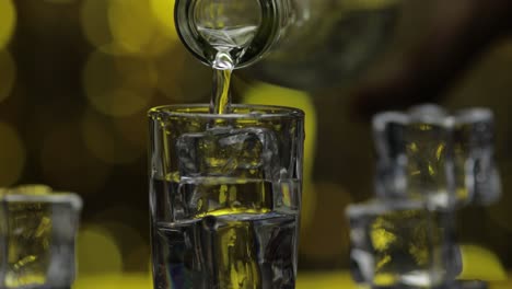 barman pour frozen vodka from bottle into shot glass. ice cubes against shiny gold party background