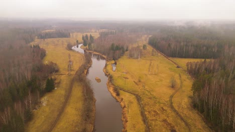 aerial view of a winding river through autumn fields and forests under an overcast sky, with visible power lines crossing the open land.