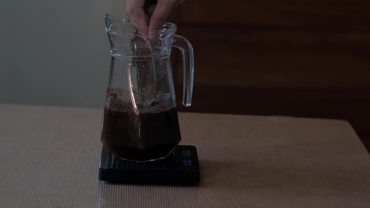 Inquadratura statica media di un cucchiaio che mescola caffè macinato e acqua fredda in una brocca di vetro durante la preparazione casalinga del cold brew