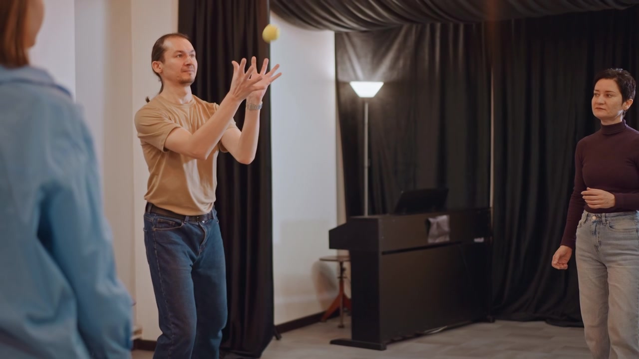 Ensemble warmup scene white performers practice near piano, tutor offers feedback, actor improvisation and timing drills, ball passing exercise for rhythm, breathing and presence, studio lights