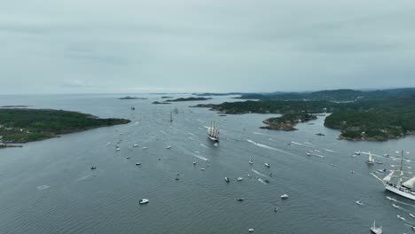 numerous sailing vessels move through the calm waters, surrounded by lush green islands under a gray sky. a vibrant maritime scene showcases the beauty of norway's coastline