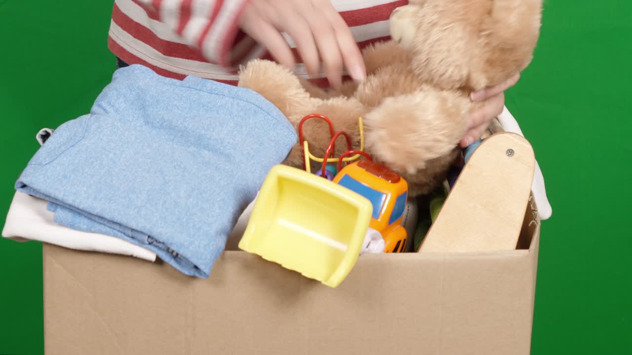 자선 기부를 위해 옷과 장난감을 준비하는 여성. 여성 손이 어린이 장난감을 분류하고 포장하고, 어린이 옷을 녹색 화면 위의 카드보드 상자에 넣습니다.