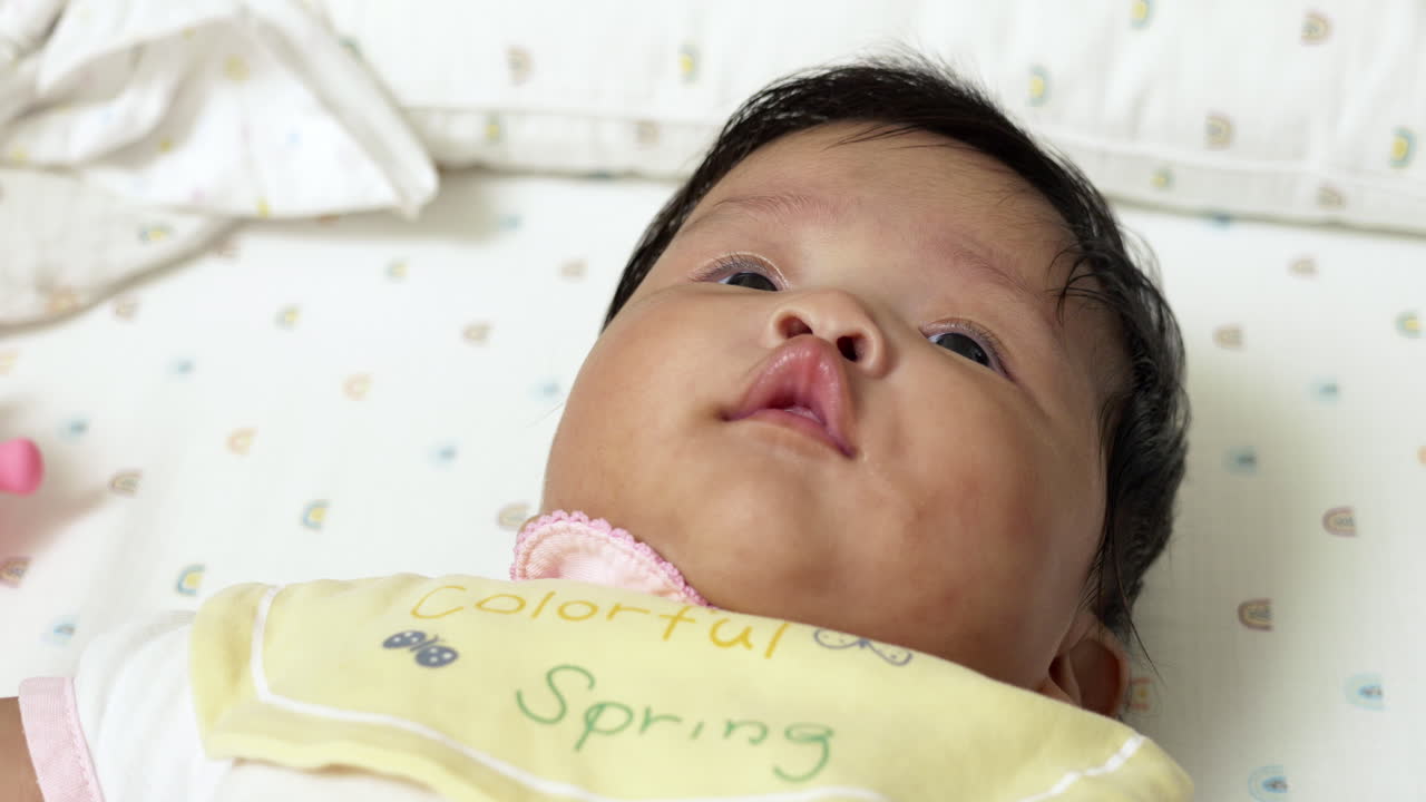 Asian newborn girl, contentedly lying on a bed with drool, expressing joy and early life development. Ideal for illustrating tender moments and baby care.