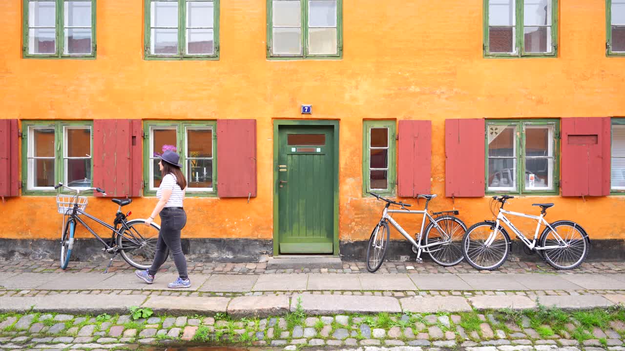 Kvinde går foran Nyboders farverige gule hus med parkerede cykler på brostensbelagt gade, København