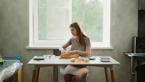 wide shot of young lady seated at chair cutting loaf of bread on table next to bowl of eggs and plates in modern kitchen with speaker in corner, natural light shining through window onto counter
