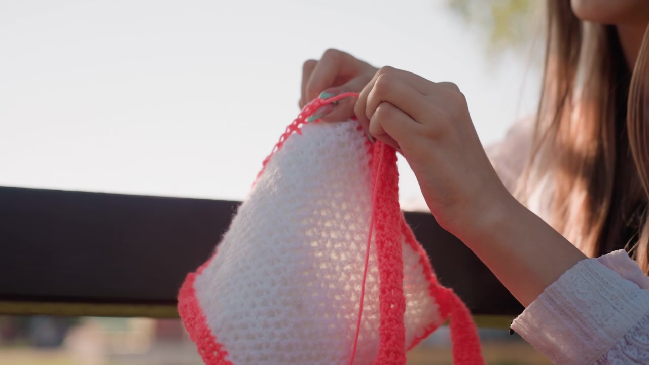 Close up of young woman crocheting with pink yarn outdoors on wooden bench. Soft natural light highlights hands and red yarn against blurred greenery background. Crochet hook in use fabric