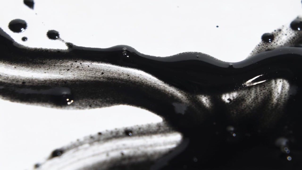 Spreading black ink stroke thickening, initiating glossy right pool on white surface with droplets