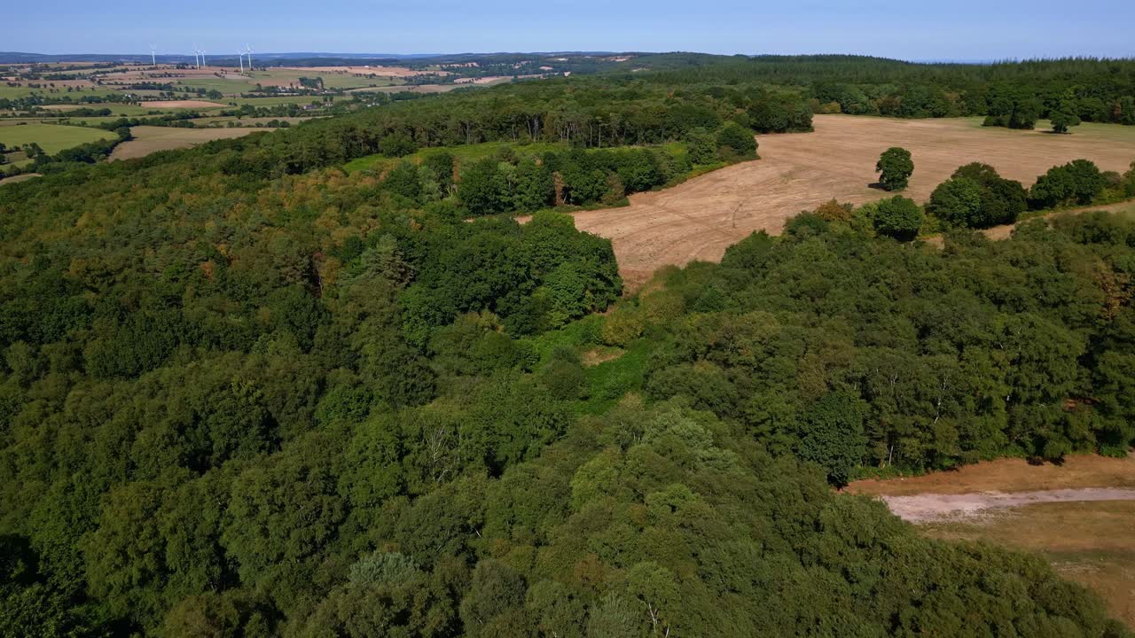 Rural landscape in Mayenne, France, lush green forest, fields, and wind turbines on horizon. Aerial drone forward