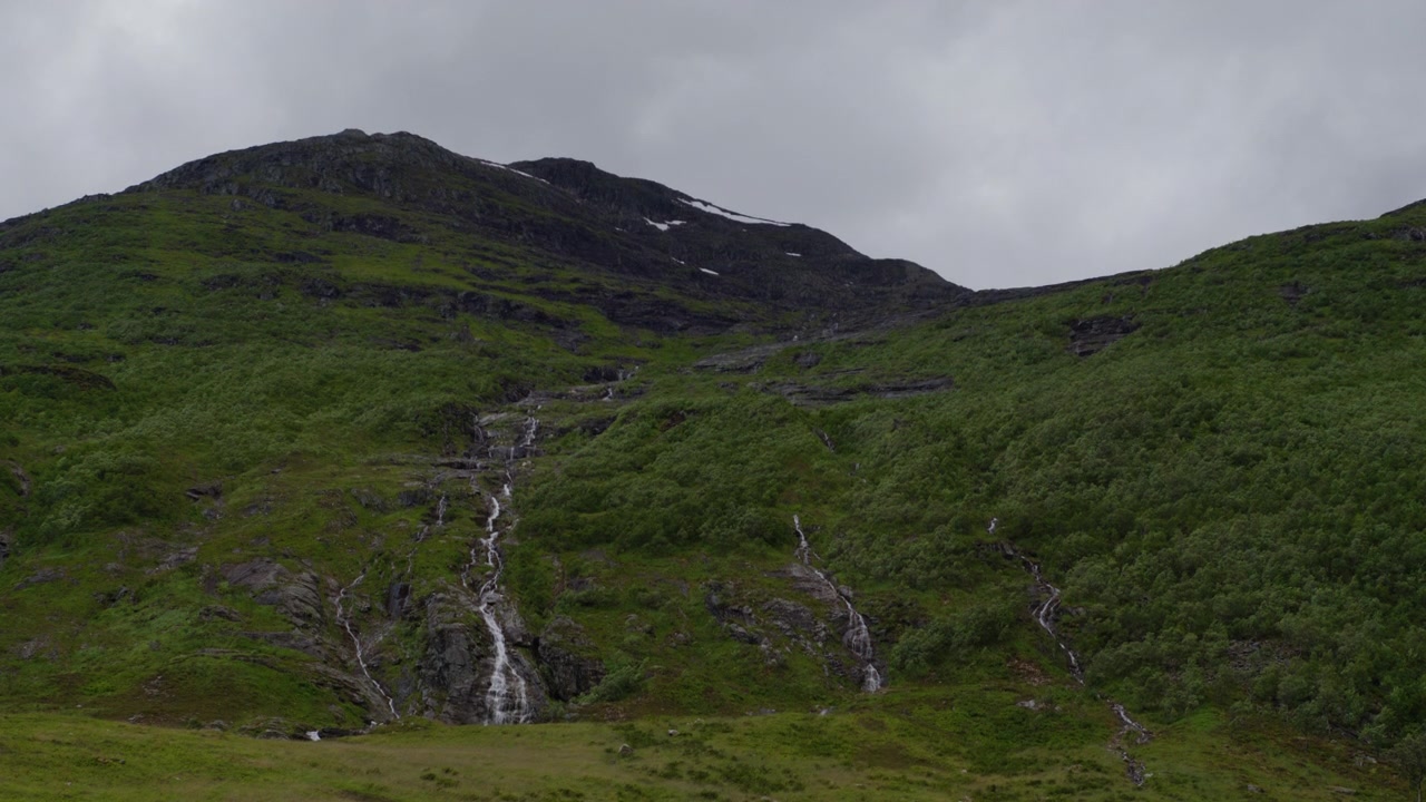 Tynne fossefall sildrer ned en bratt, steinete åsside dekket av lav vegetasjon under en grå himmel i Vest-Norge.